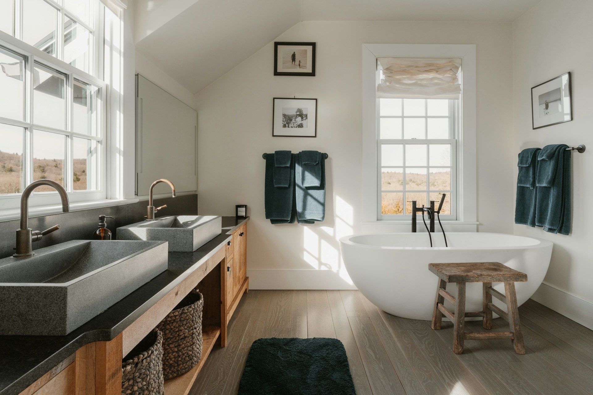 Modern bathroom with dual stone sinks, wood vanity, freestanding tub, teal towels, and windows overlooking a landscape.